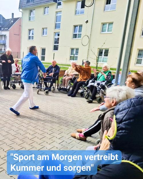Eine Aufnahme im Freien zeigt einen gepflasterten Bereich, in dem eine Gruppe älterer Menschen an einer Form von Bewegung oder Aktivität teilnimmt, die von einem jüngeren Mann geleitet wird.  Die älteren Erwachsenen sind entweder in Rollstühlen oder auf Stühlen sitzend, wobei einige aktiv teilnehmen, während andere zuschauen. Der Mann, der die Gruppe anführt, trägt eine leuchtend blaue Jacke.  Die Gruppe befindet sich vor einer Pflegeeinrichtung, im Hintergrund ist ein sichtbarer Teil des Gebäudes zu erkennen.  Ein deutscher Satz, der am unteren Rand des Bildes steht, lautet: „Sport am Morgen, vertreibt Kummer und Sorgen“